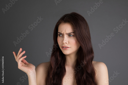 Young brunette woman with bare shoulders smirking from disbelief frowning while pointing at apper left corner