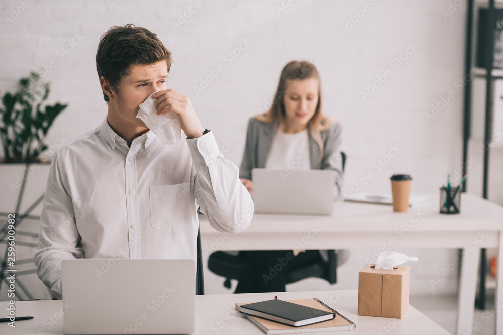 selective focus of businessman sneezing in tissue while using laptop near businesswoman in office selective focus of businessman sneezing in tissue while using laptop near businesswoman in office