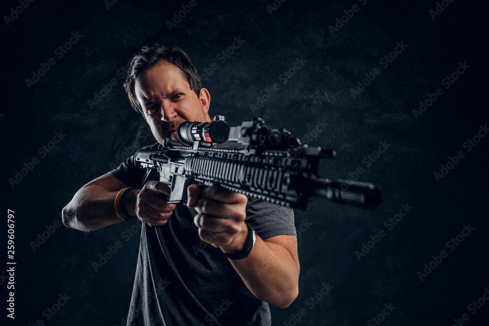 Distraught middle-aged man dressed in casual clothes holding an assault rifle and aims at the target. Studio photo against a dark textured wall