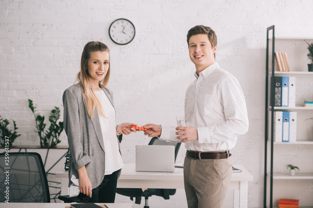 Obraz premium happy businessman holding pills and glass of water near cheerful blonde colleague in office