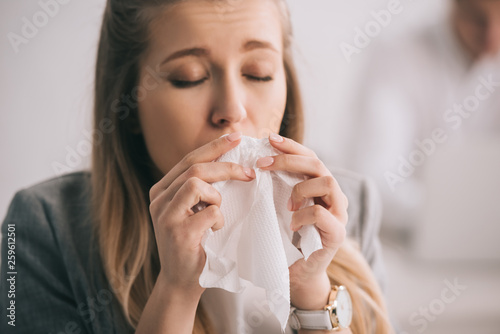 blonde businesswoman sneezing in tissue with closed eyes in office