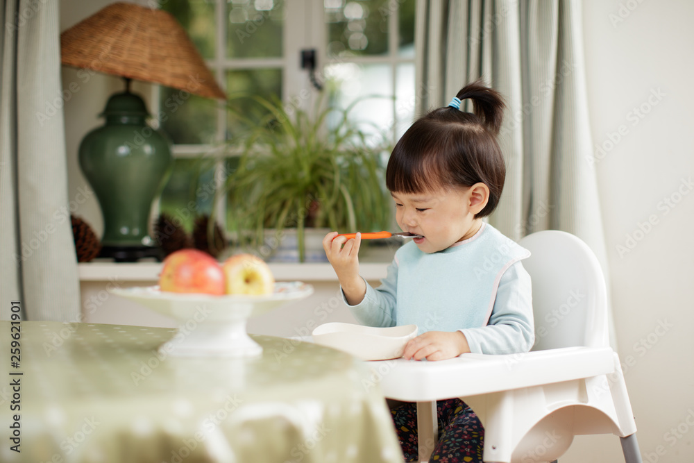 toddler girl eating healthy vegetable sitting on high chair beside a dinner table at home toddler girl eating healthy vegetable sitting on high chair beside a dinner table at home