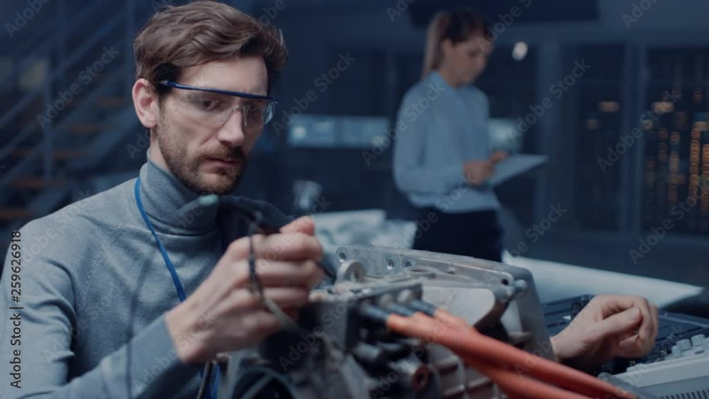 Professional Automotive Engineer in Glasses with a Computer and Inspection Tools is Testing an Used Electric Engine in a High Tech Laboratory with a Concept Car Chassis. Professional Automotive Engineer in Glasses with a Computer and Inspection Tools is Testing an Used Electric Engine in a High Tech Laboratory with a Concept Car Chassis.