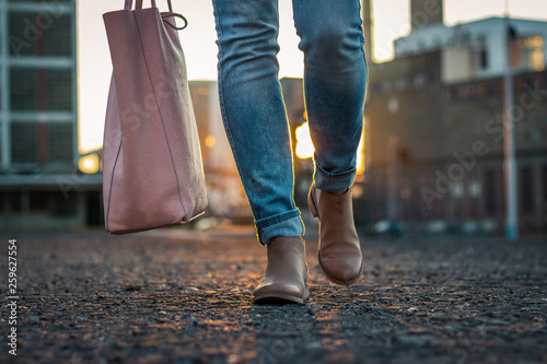 Woman in jeans and stylish leather shoe with bag walking in city during sunset