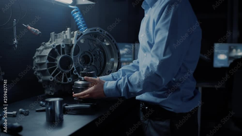 Professional Automotive Engineer in Glasses is Working on Transmission Gears in a High Tech Innovative Laboratory with a Computer Screens.