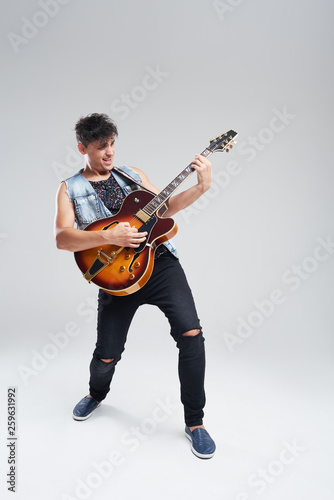 Young man musician with an electric guitar in hand on a gray background. he plays rock and roll loudly. Full-length portrait.