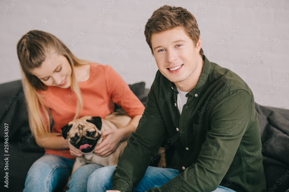selective focus of handsome smiling man sitting near woman with adorable pug dog