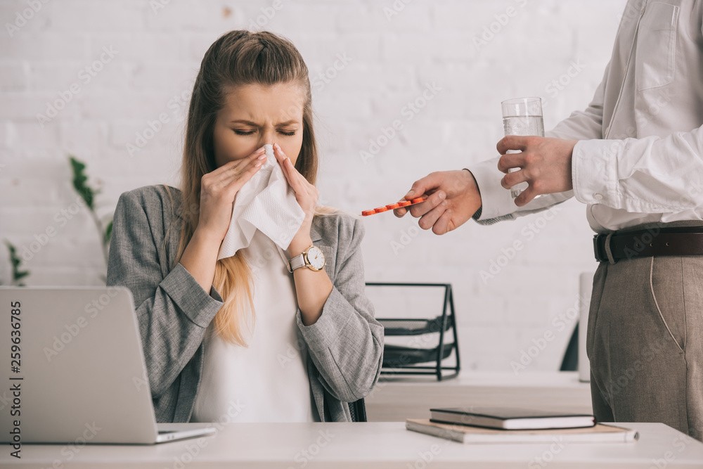 blonde businesswoman sneezing in tissue with closed eyes near coworker holding pills and glass of water