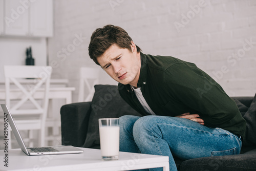Fotografie upset man looking at glass of milk while holding stomach and sitting on sofa