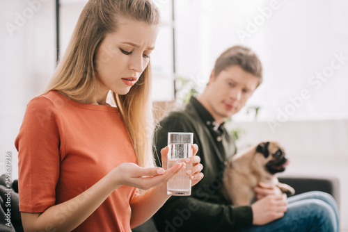 selective focus of upset blonde woman allergic to dog holding pills and glass of water near man with pug