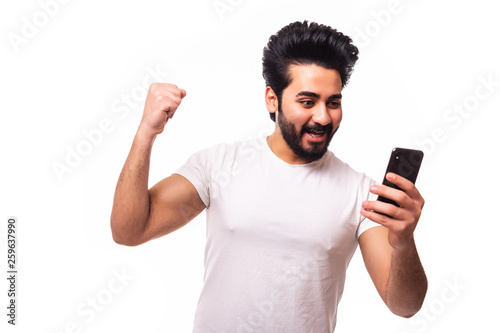 Portrait of an excited young arab man celebrating looking at mobile phone isolated over white background