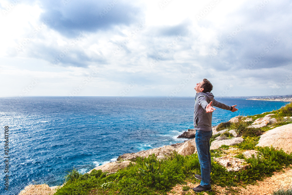 Rear view of a young guy standing on a seaside rock cliff edge with arms spread open against a seascape