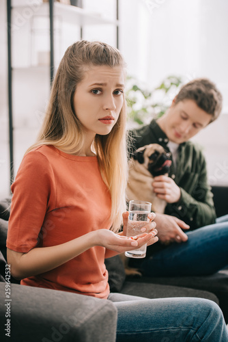 selective focus of upset girl allergic to dog holding pills and glass of water near man with pug at home