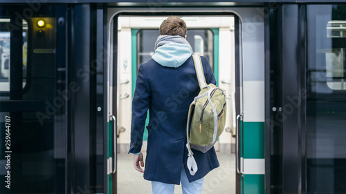 Close up rear view of handsome man in blue coat and green backpack behind stands in front of the open doors of the subway and waiting for next train. Way to work. Urban life concept.