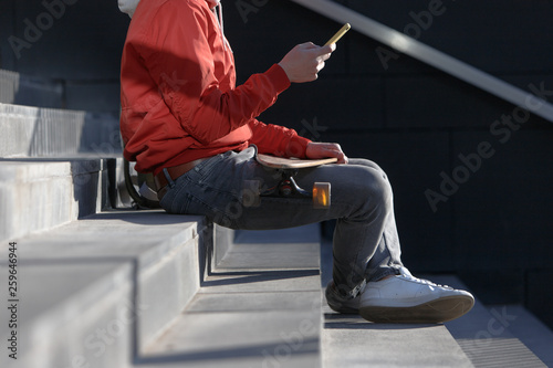 Stylish man longboarder in casual clothes using his smartphone, resting on the steps, sitting with longboard/skateboard outdoors, top view, cropped image. Urban, subculture, skateboarding concept