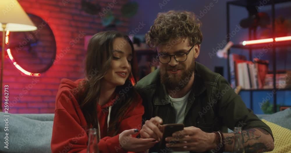 Close up of the young happy Caucasian boyfriend and girlfriend sitting on the sofa at night and looking at the smartphone screen while talking. Close up of the young happy Caucasian boyfriend and girlfriend sitting on the sofa at night and looking at the smartphone screen while talking.
