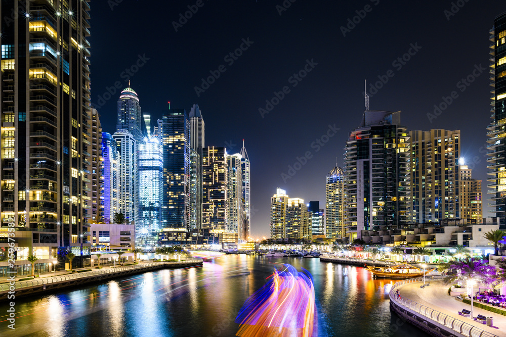 Stunning view of the Dubai Marina at dusk with illuminated skyscrapers in the background and a light trails left by a yacht sailing in the foreground. Dubai, United Arab, Emirates.