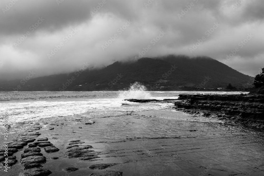 Waves crash and ripple onto the rocky shoreline under a moody, misty, rainy sky.