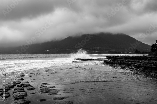 Waves crash and ripple onto the rocky shoreline under a moody, misty, rainy sky.