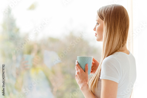 Close-up profile side view portrait of nice cute charming attractive peaceful calm dreamy straight-haired girl wearing white tshirt holding in hands cup looking at window in light white interior room