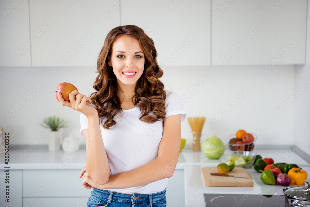 Portrait of her she nice-looking attractive lovely cute charming winsome cheerful cheery wavy-haired girl holding in hand apple in modern light white interior style room