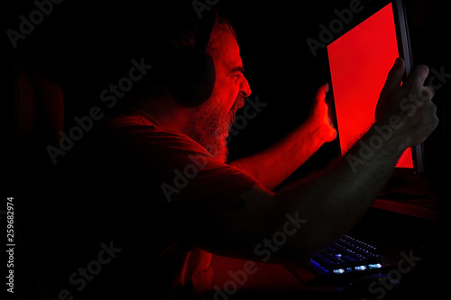 Angry bearded man in red light screaming in rage at PC monitor and shaking it violently 