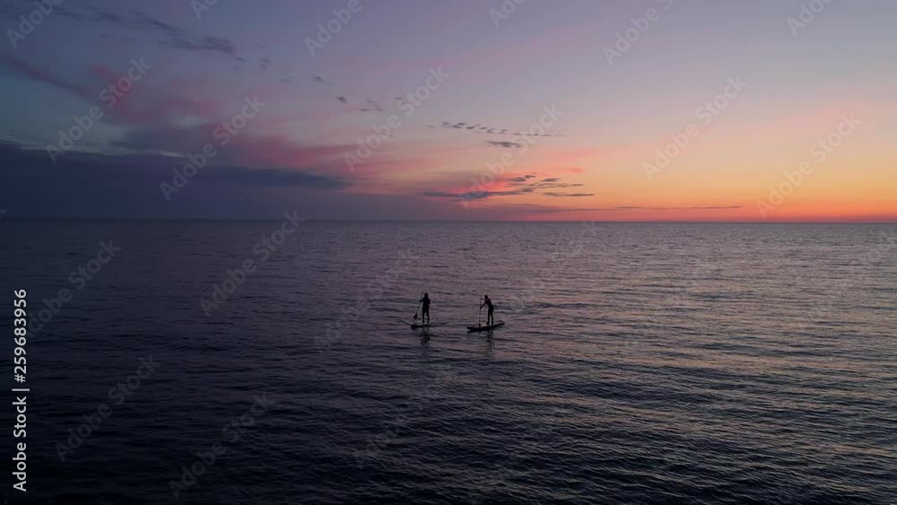 Aerial view couple silhouettes on paddle boards in sea during sunset. Amazing orange sky in background.