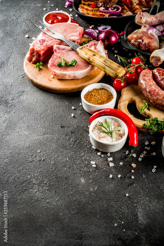 Various raw meat ready for grill and bbq, with vegetables, greens, sauces kitchen grilling utensils. Chicken legs, pork steaks, sausages, beef ribs with herbs, black concrete background copy space