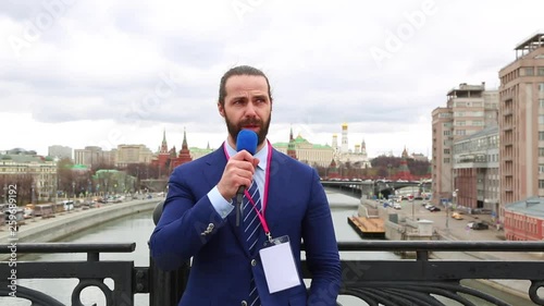 Male reporter with a microphone tells news camera on the background of the city.