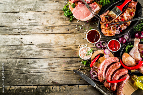 Various raw meat ready for grill and bbq, with vegetables, greens, sauces kitchen grilling utensils. Chicken legs, pork steaks, sausages, beef ribs with herbs, rustic wooden background copy space