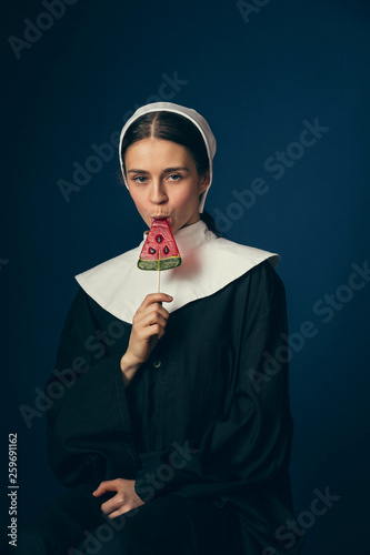 New capabilities. Medieval young woman as a nun in vintage clothing and white mutch sitting on the chair and eating sweets on dark blue background. Concept of comparison of eras. photo