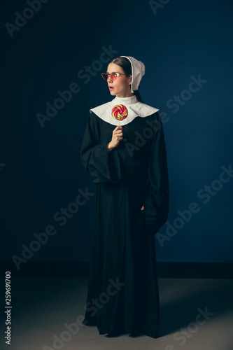 Scare of expose. Medieval young woman as a nun in vintage clothing, white mutch and red sunglasses eating sweets and standing on dark blue background. Concept of comparison of eras. photo