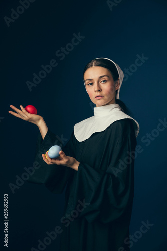 Ideal combination. Medieval young woman as a nun in vintage clothing and white mutch standing with the Easter colored red, yellow eggs and on dark blue background. Concept of comparison of eras. photo