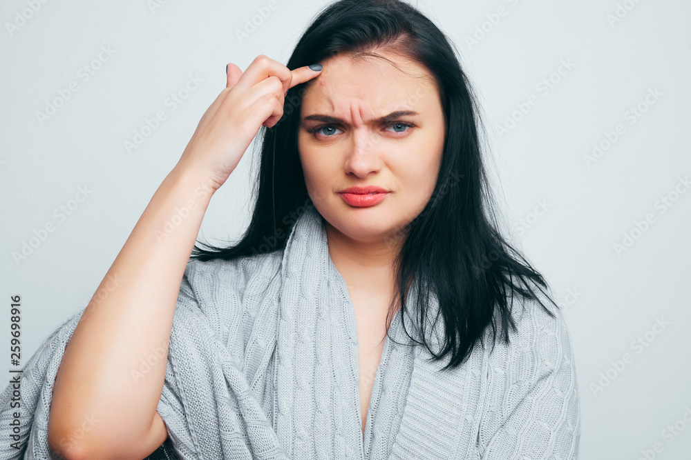 Upset girl in a gray robe indicates poor skin, age wrinkles, acne on a white and gray studio background. A woman is looking for flaws and age changes.
