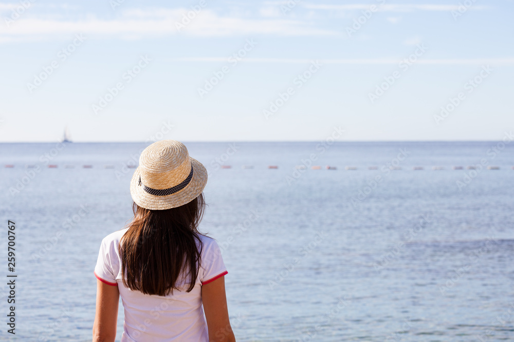 Beautiful girl looks at the sea. Young girl in a hat looking at a calm sea and blue skies back view.