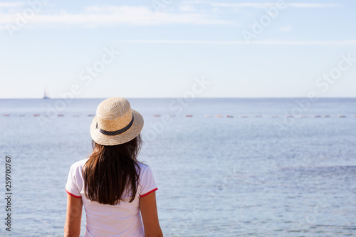 Beautiful girl looks at the sea. Young girl in a hat looking at a calm sea and blue skies back view. © Nickolay Khoroshkov