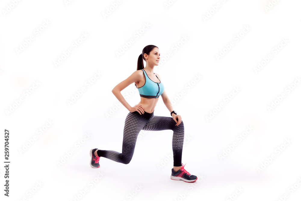 Fototapeta premium A dark-haired woman coach in a sporty short top and gym leggings makes lunges by the feet forward, hands are held out to the side on a white isolated background in studio