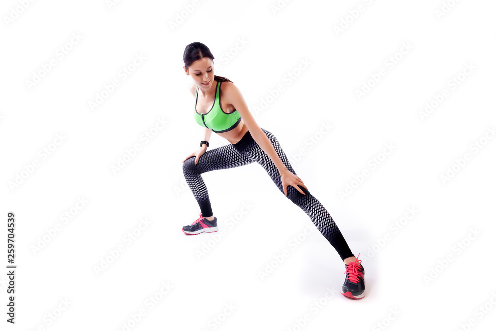 A dark-haired woman coach in a sporty  short top and gym leggings makes lunges  by the feet side, hands are held out to the side   on a  white isolated background in studio