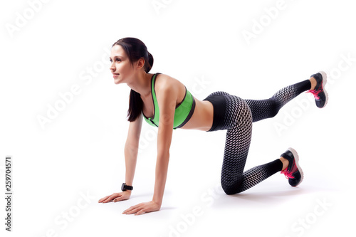 A dark-haired woman coach in a sporty short top and gym makes and stretch legs, doing exercise on a white isolated background in studio. Fit girl living an active lifestyle