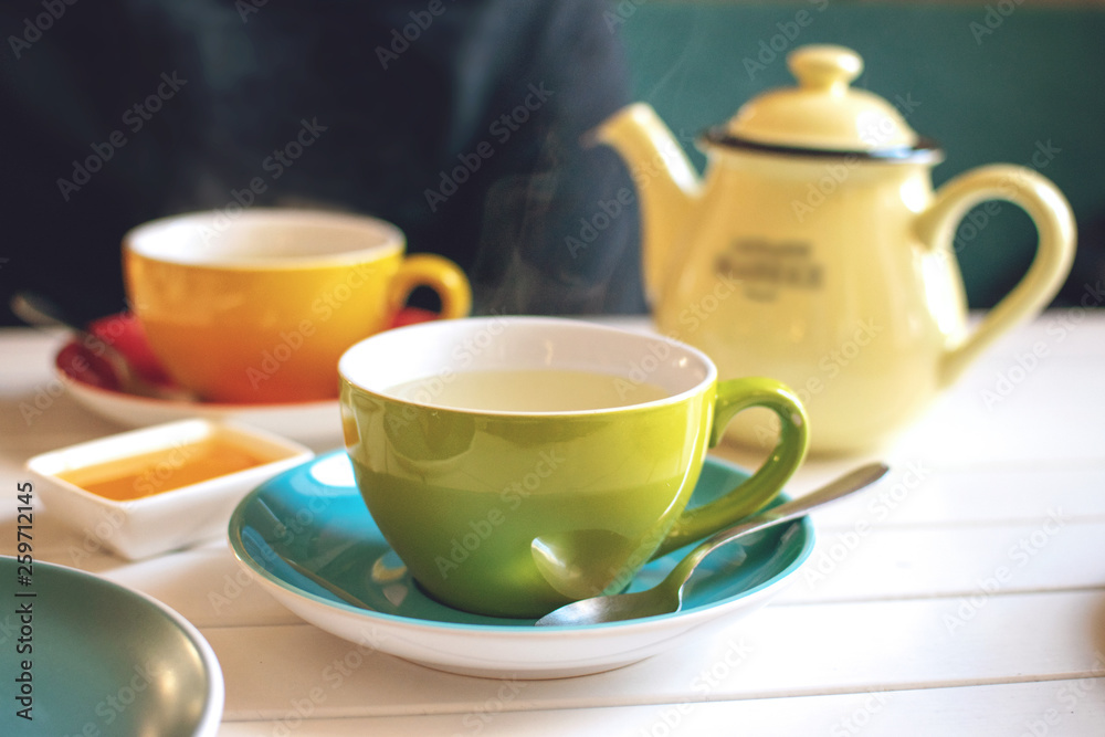 Herbal tea in green cup, honey and yellow teapot on white wooden table in cafe. A cup of hot tea with steam. Natural light Herbal tea in green cup, honey and yellow teapot on white wooden table in cafe. A cup of hot tea with steam. Natural light