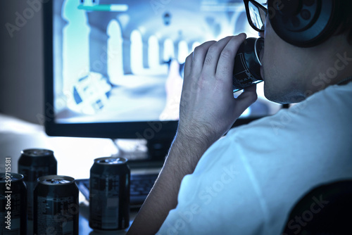 Guy drinking soda and playing video game or watching online live stream. Too much energy drink. Many empty cans of cola. Addicted gamer and computer. Sugar addiction. Geek and nerd with glasses.