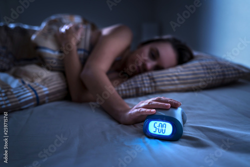 Tired woman waking up for work or school early in the morning. Grumpy lady pushing snooze button or turning off alarm clock with hand. Getting up after sleeping. Exhausted person in bed. Dark bedroom.
