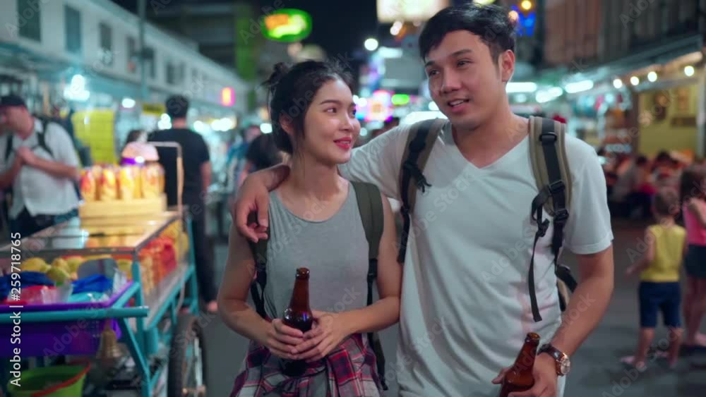 Slow motion - Traveler backpacker Asian couple travel in Bangkok, Thailand, sweet couple drinking alcohol or beer while hangout at The Khao San Road. Couple travel in Thailand concept. Slow motion - Traveler backpacker Asian couple travel in Bangkok, Thailand, sweet couple drinking alcohol or beer while hangout at The Khao San Road. Couple travel in Thailand concept.