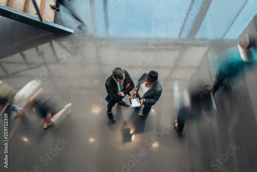 Two business people standing in the lobby of an office looking at a tablet while people are walking past in a blur