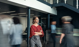 Beautiful young business woman standing in a busy lobby of an office with her arms crossed, looking ahead, with people walking past in a blur