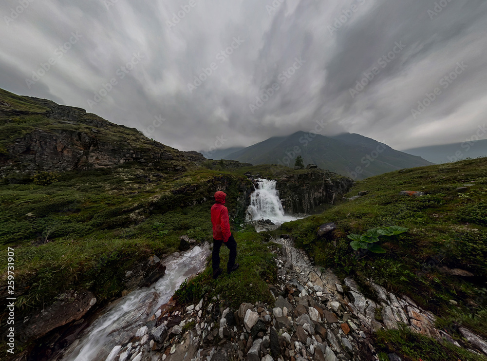 man in red stands under waterfall on black rock in mountains on cloudy rainy day.. Wide panorama