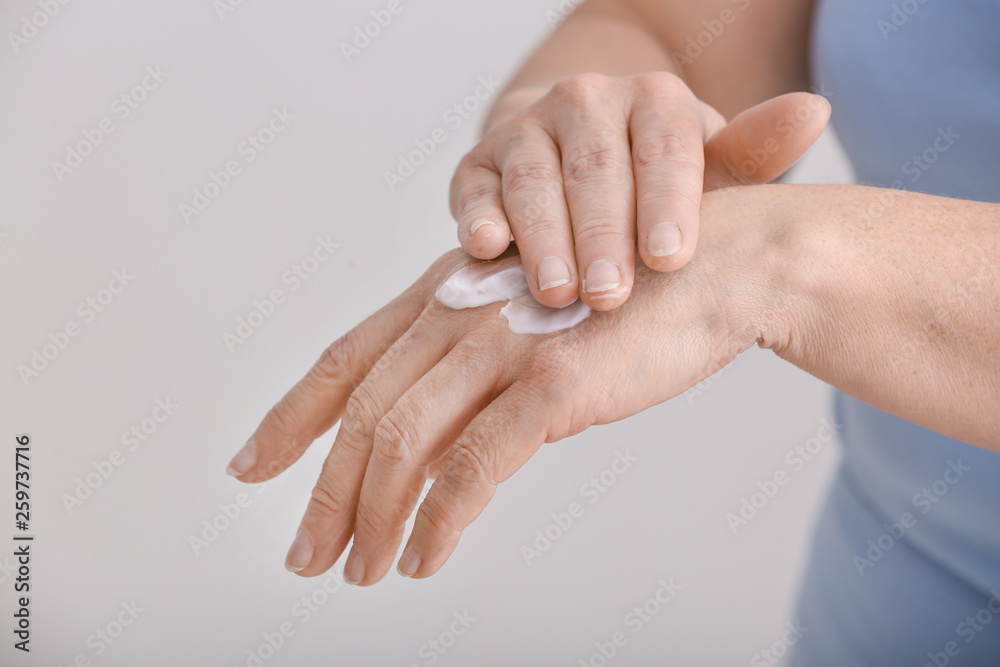 Mature woman applying hand cream on grey background, closeup