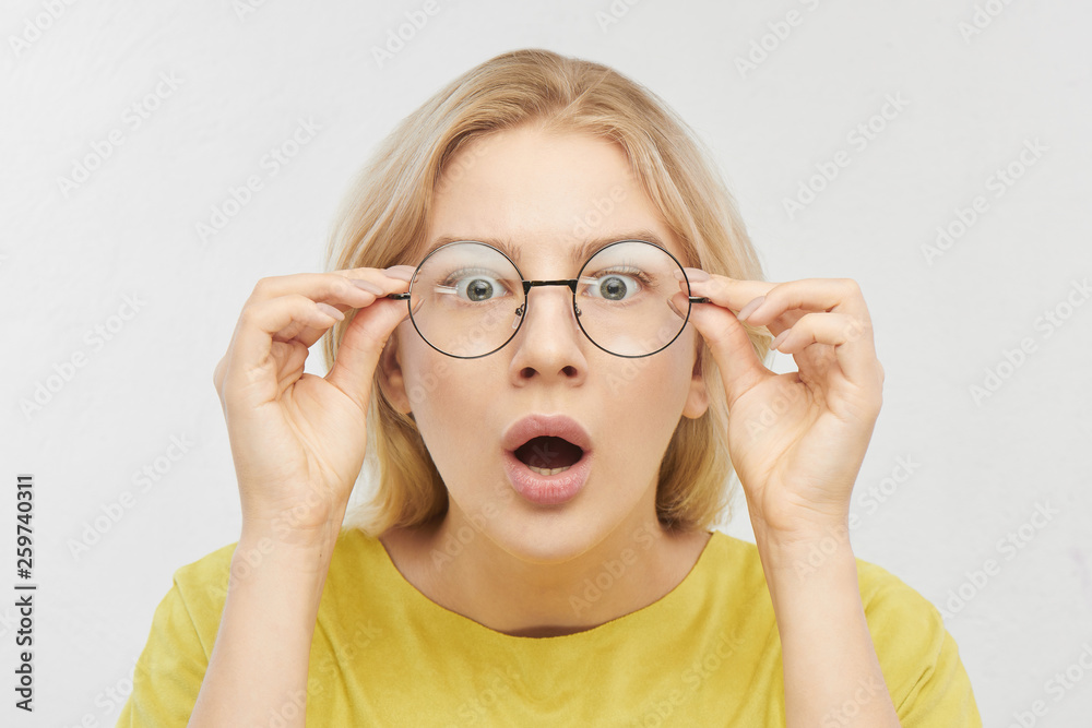 Surprised woman in round glasses with open mouth and bulging eyes looks into camera and sees something incredible and amazing isolated on white background