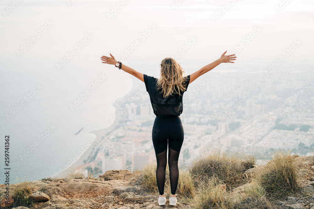 Sporty young woman enjoying freedom with open hands on the top of mountain looking the city from above Sporty young woman enjoying freedom with open hands on the top of mountain looking the city from above