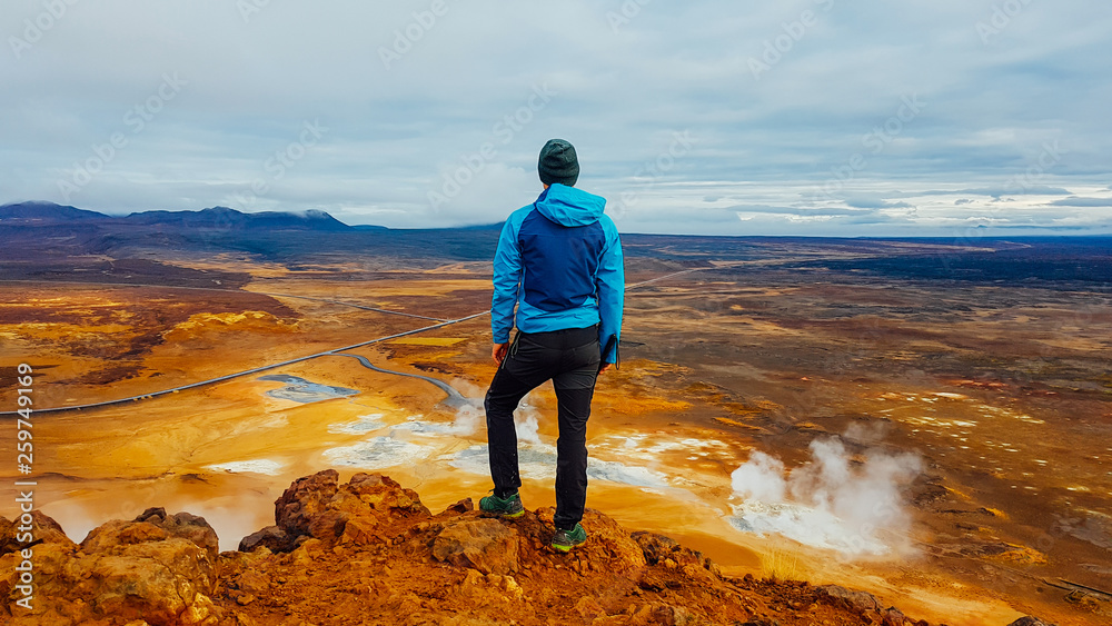 Obraz premium Young man wearing a blue jacket stands on top of a mud mountain, overlooking a geothermal spot noted for its bubbling pools of mud & steaming fumaroles emitting sulfuric gas. Power of planet Earth.